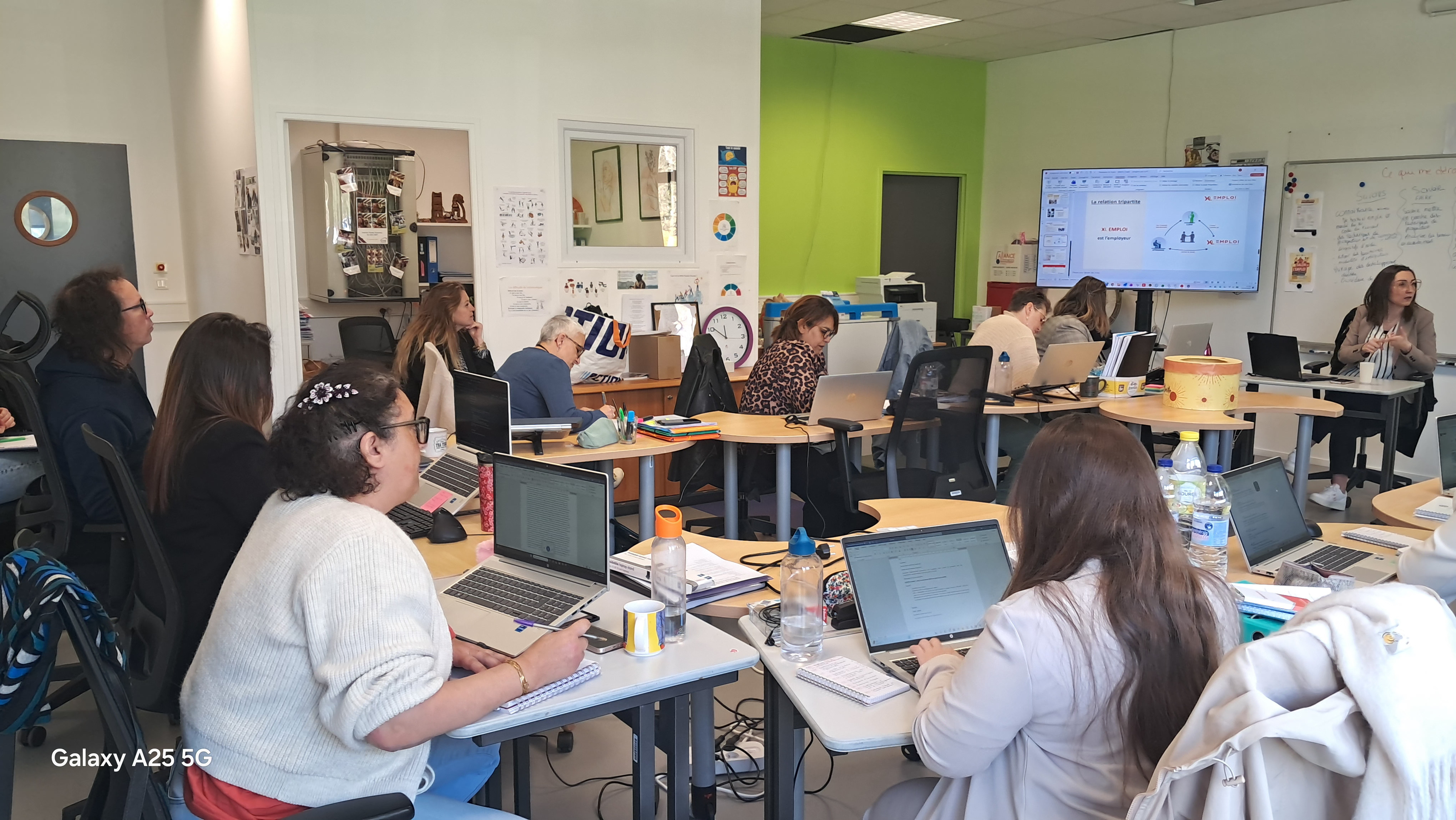 Salle de formation : des adultes travaillent sur ordinateurs tandis qu’une formatrice présente devant un écran et un tableau blanc.
