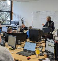une personne debout dans une salle de classe avec des ordinateurs et des personnes qui l'écoute
