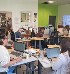 Salle de formation : des adultes travaillent sur ordinateurs tandis qu’une formatrice présente devant un écran et un tableau blanc.